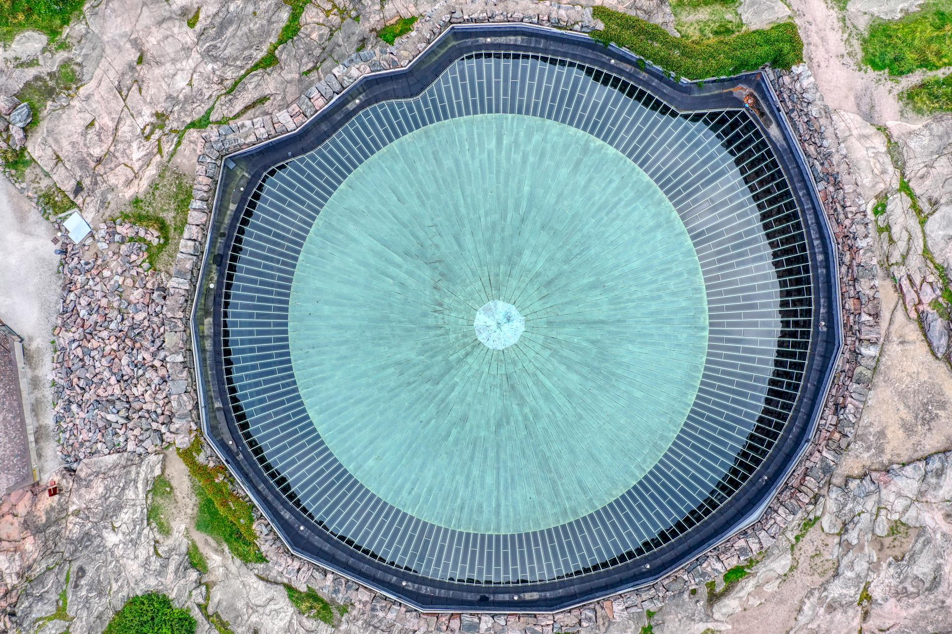 Ilmakuva Temppeliaukion kirkosta. Kuvan keskellä on kirkon vihertävä kuparikatto, jota kiertävät lasiset ikkunat. Kirkkoa ympäröi louhoskivimuuri. Aerial picture of Temppeliaukio Church. The green copper roof of the church is surrounded by skylight windows and a quarry wall.